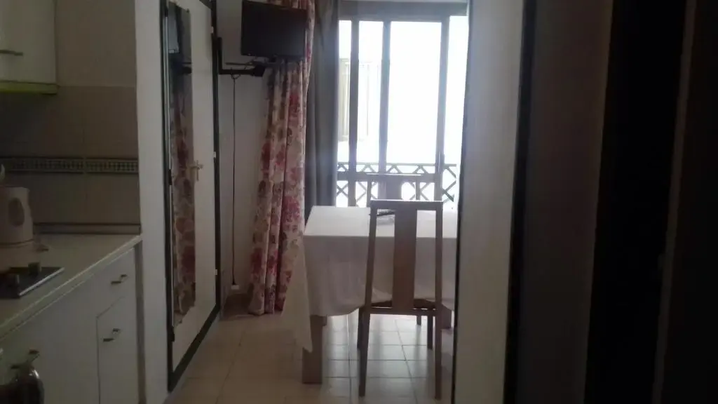 Cocina y mesa con mantel blanco junto a un balcón en un estudio, con televisor en la pared