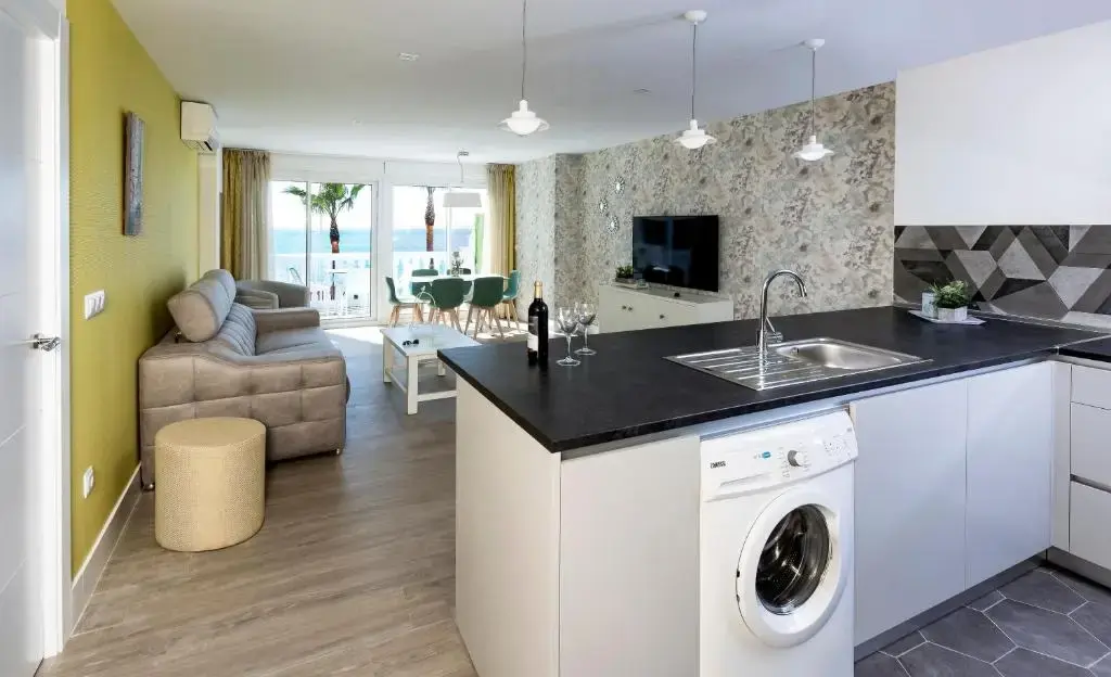 Salón con sofá, cocina americana con lavadora y comedor junto al balcón con vistas al mar