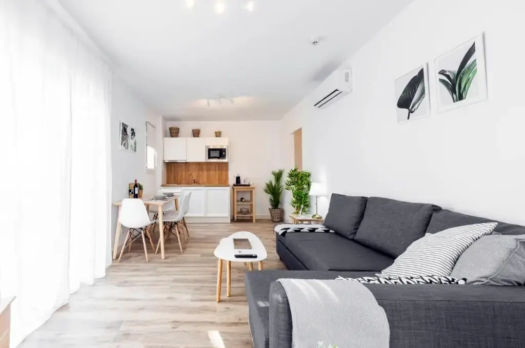 Salón con sofá rinconera gris abierto a la cocina blanca con microondas y mesa de comedor