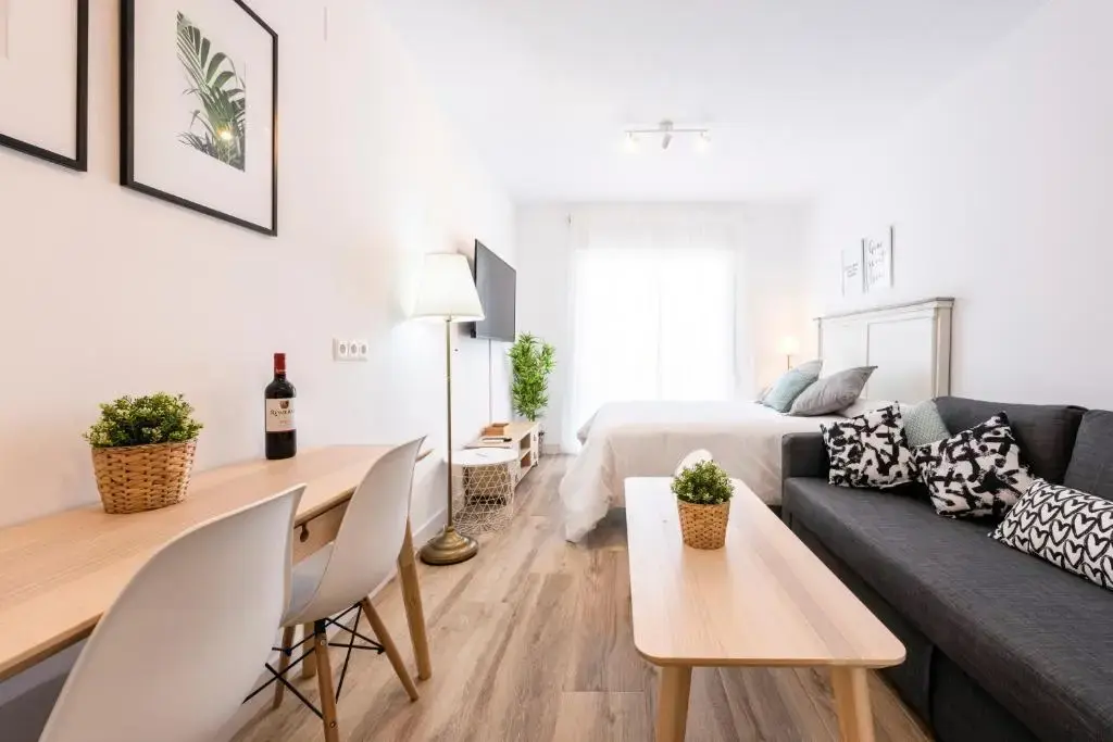 Estudio con cama de matrimonio, sofá gris, mesa auxiliar y escritorio con dos sillas blancas
