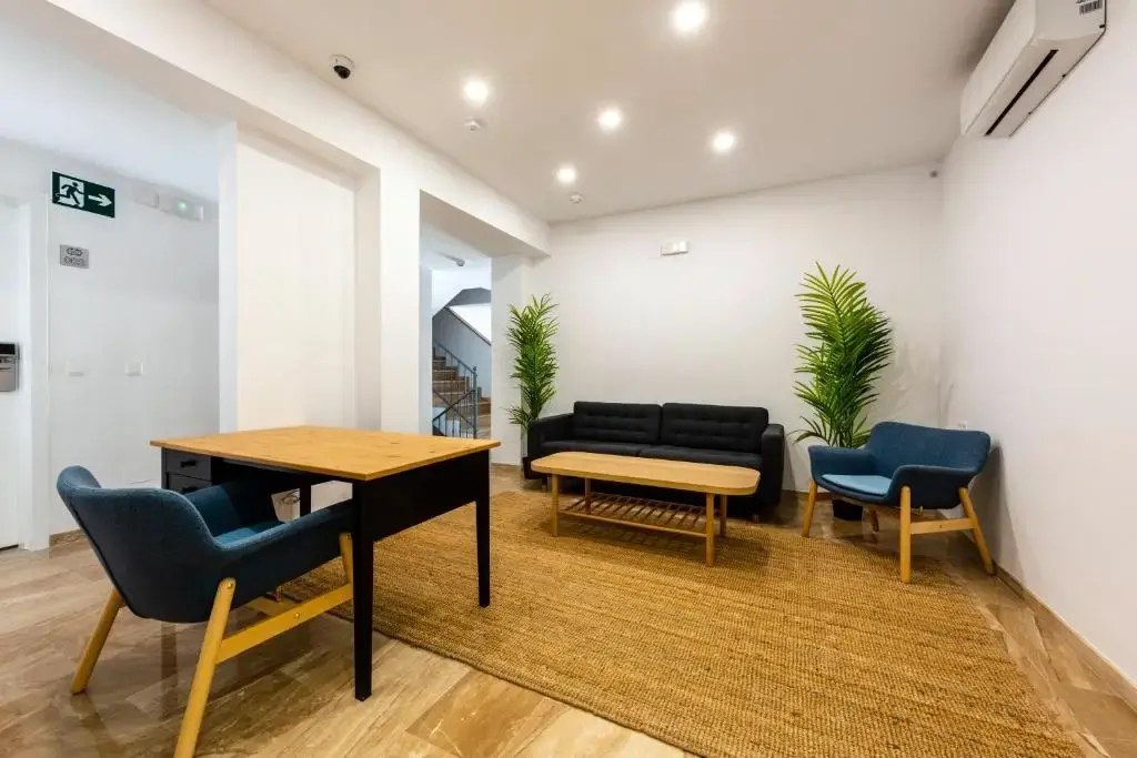 Zona común de entrada con escritorio, sofá negro, sillones azules y alfombra de fibra