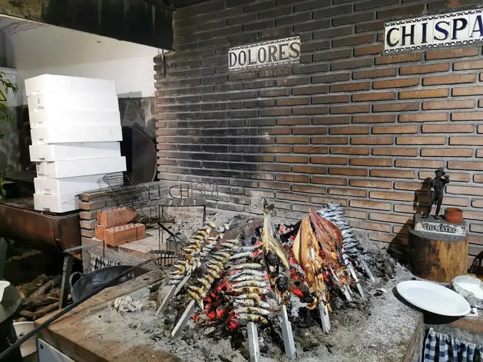 Asador de leña con espetos de sardinas y pescados junto a la pared de ladrillo