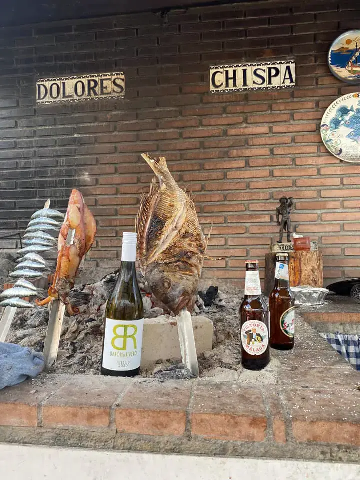 Pescado, cigala y sardinas asándose en espetos junto a botellas de vino y cerveza