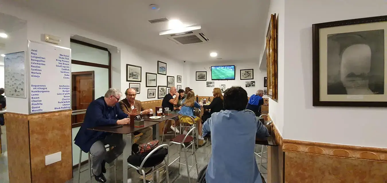 Salón interior con mesas altas, cuadros enmarcados y un televisor con fútbol