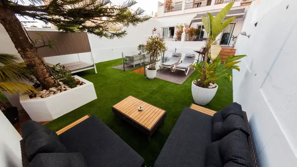 Patio con césped artificial, sofás de exterior, mesa de madera, tumbonas y plantas en macetas