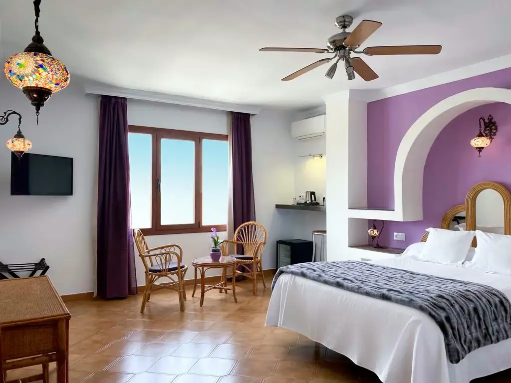Habitación amplia con pared morada en arco, lámparas de mosaico, ventilador y sillas de mimbre