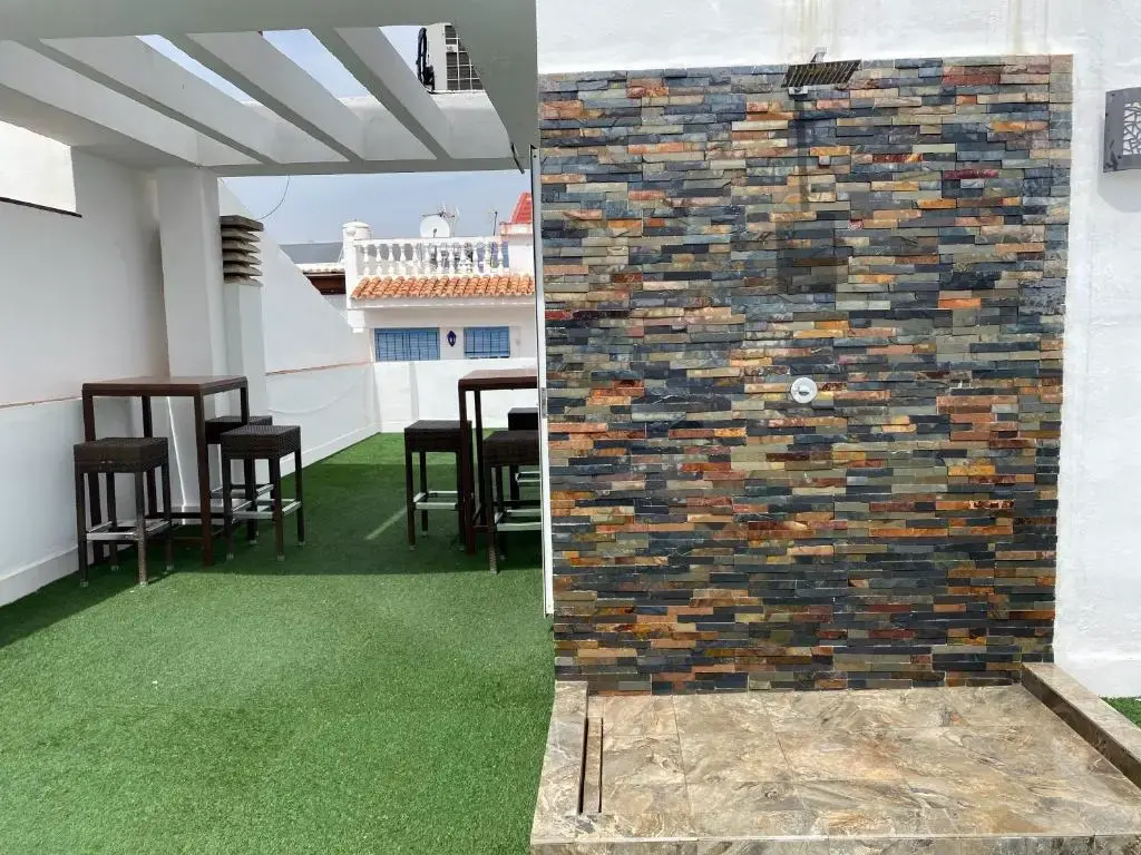 Azotea con césped artificial, mesas altas con taburetes y ducha junto a una pared de piedra