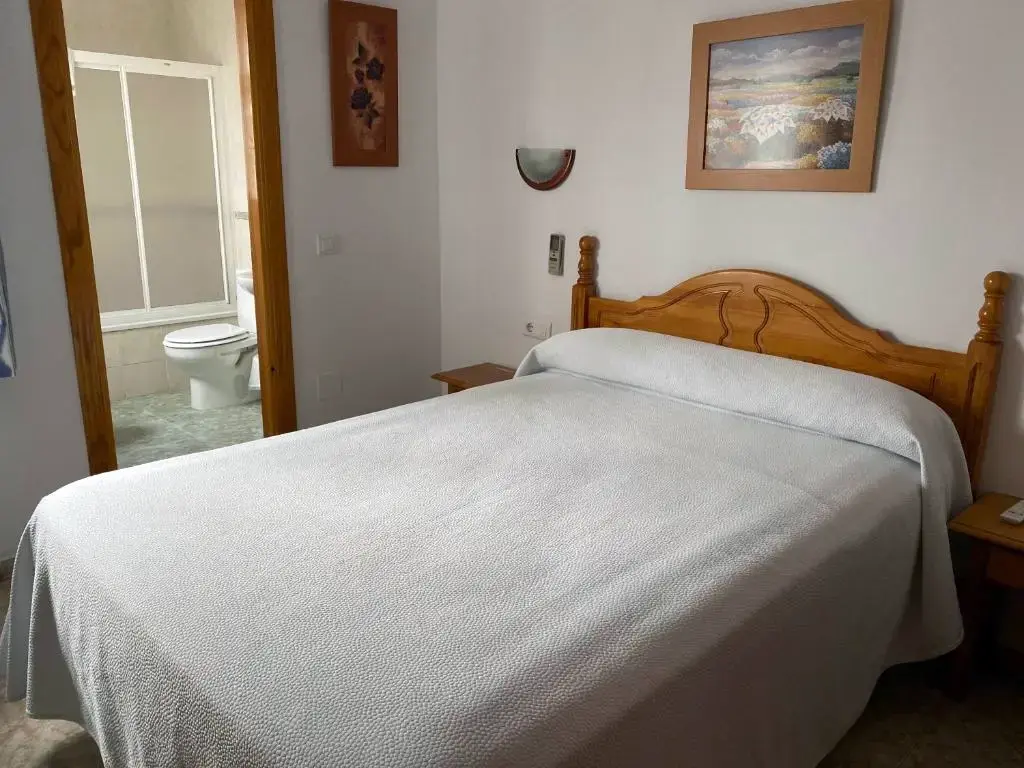 Cama de matrimonio con colcha blanca, cuadros en la pared y puerta abierta al baño