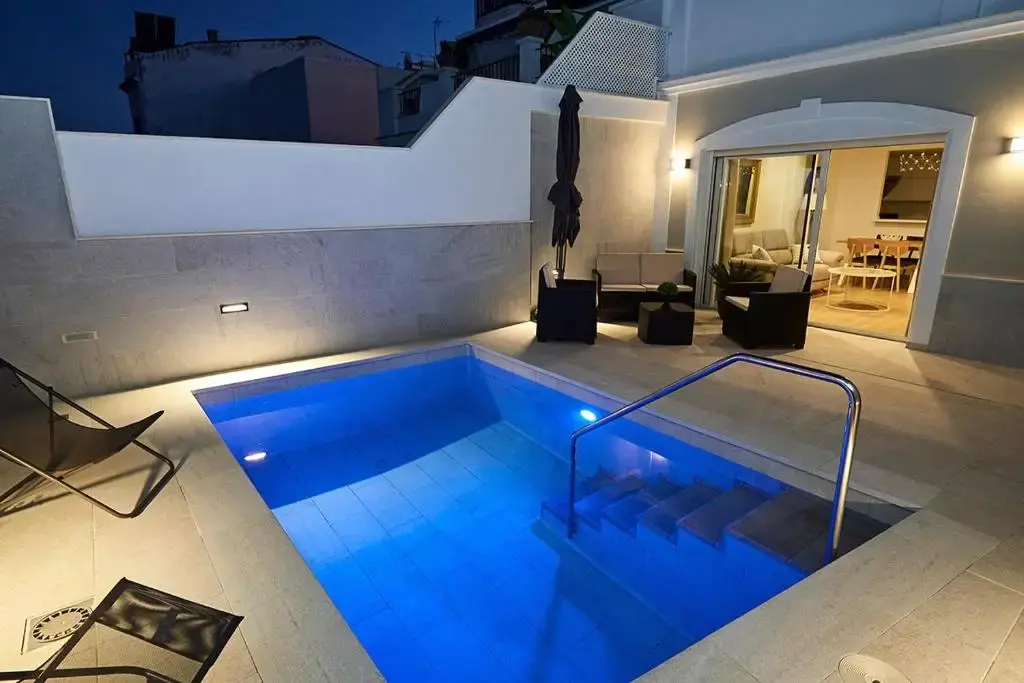 Piscina iluminada del patio al anochecer, con tumbonas y sofás junto al salón del Hostal Aurora