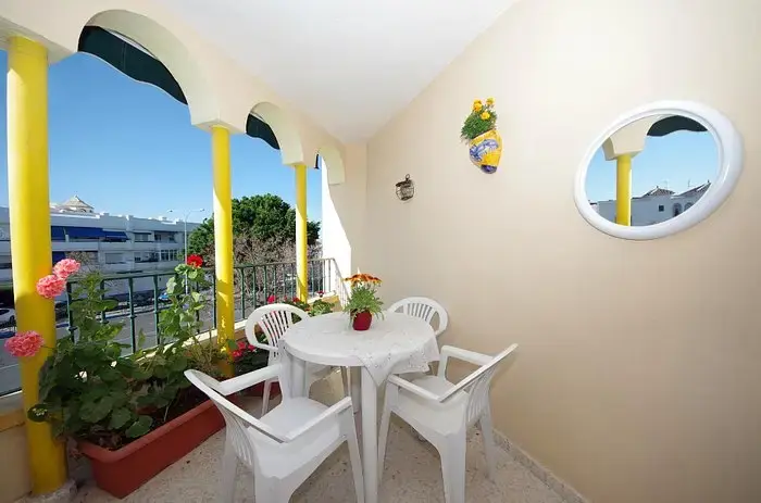 Terraza con columnas amarillas, mesa blanca, sillas y macetas con geranios sobre la avenida
