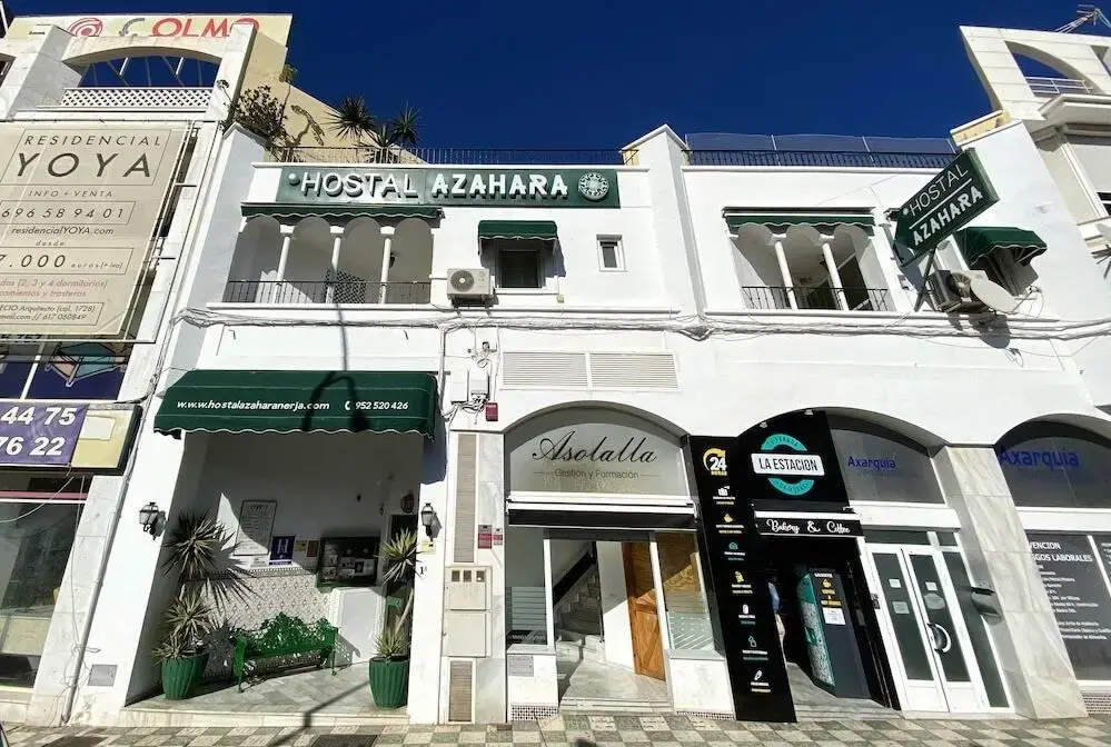 Entrada del Hostal Azahara en la avenida, entre locales comerciales y bajo un cielo azul