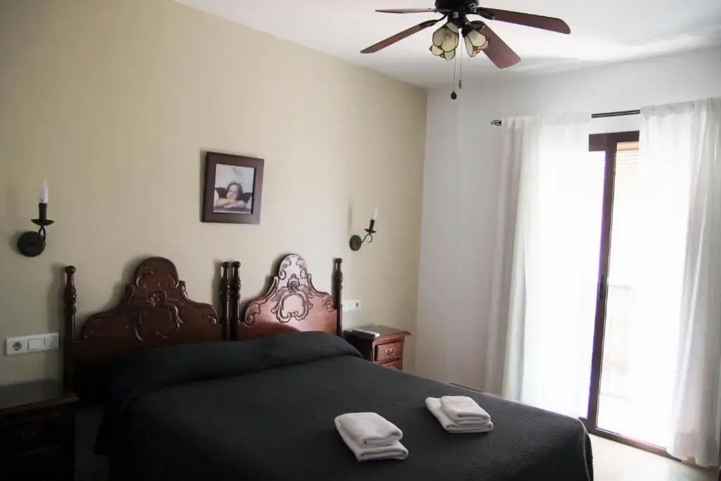 Dormitorio con cama de matrimonio de cabecero tallado, ventilador de techo y balcón con visillos