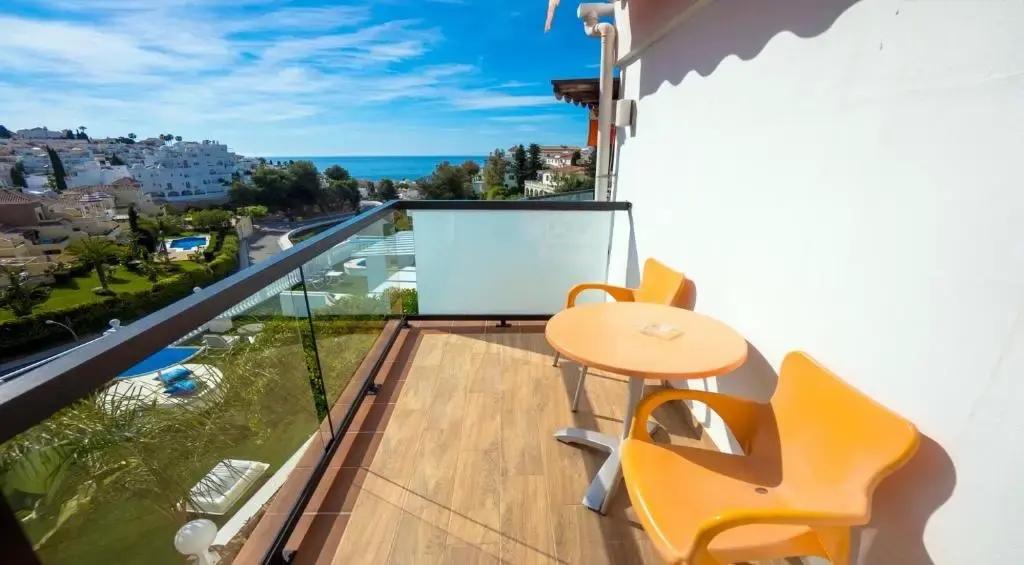 Balcón con mesa y sillas naranjas sobre jardines con piscinas y el mar en el horizonte