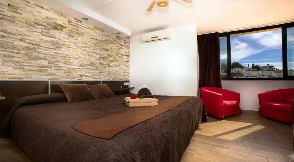 Habitación con cama grande marrón, pared de piedra, sillones rojos y ventanal con vistas
