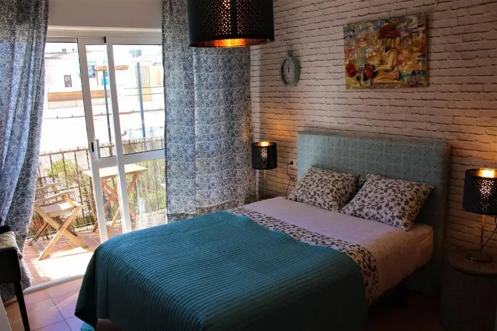 Habitación doble con colcha turquesa, pared de ladrillo blanco y balcón con sillas