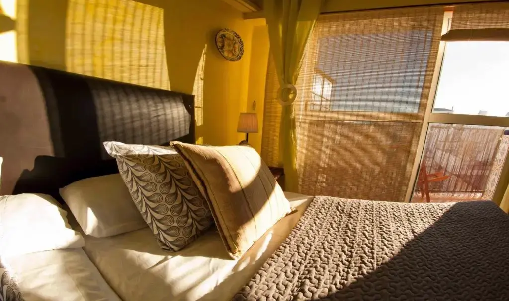Dormitorio con cama de cabecero oscuro, cojines y luz de tarde entrando por el balcón
