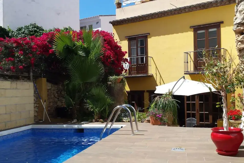 Piscina junto a la casa amarilla con balcones de forja y buganvillas rojas