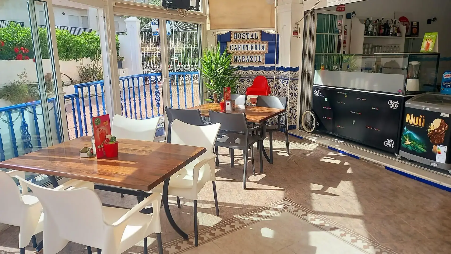 Cafetería del Marazul con mesas de madera, sillas blancas y barra con vitrina