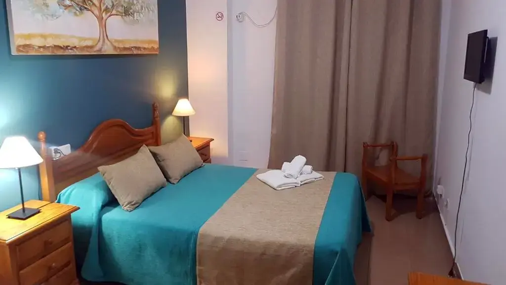 Habitación doble con colcha turquesa, cabecero de madera, toallas y televisor en la pared
