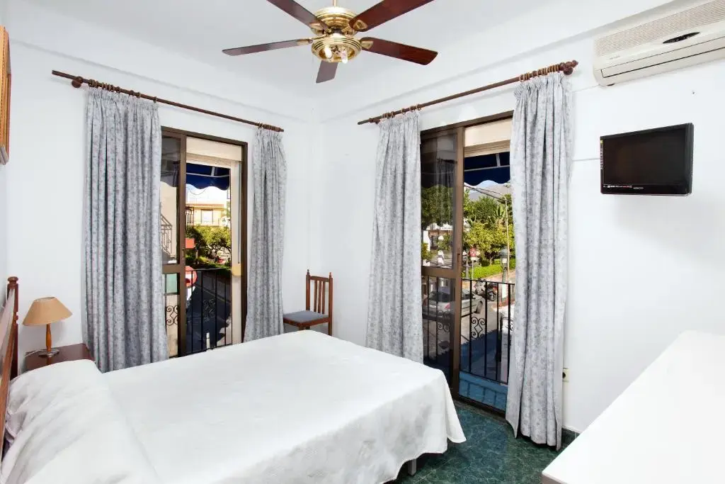 Habitación doble con ventilador de techo, televisor y dos balcones con cortinas grises