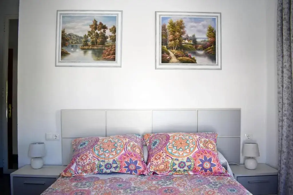 Cama de matrimonio con colcha de mandalas de colores, mesitas blancas y dos cuadros de paisaje