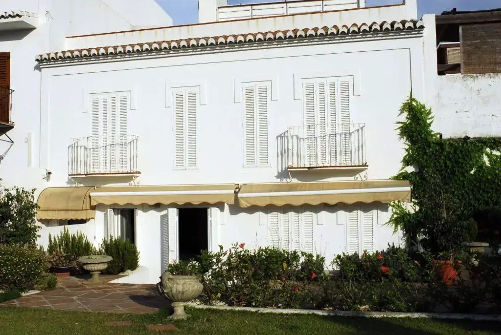 Casa blanca con contraventanas, toldos y balcones de forja frente a un jardín con flores