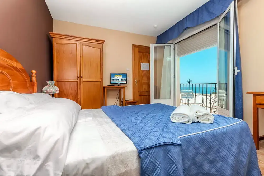 Habitación con cama de colcha azul, televisor y balcón abierto con vistas al mar