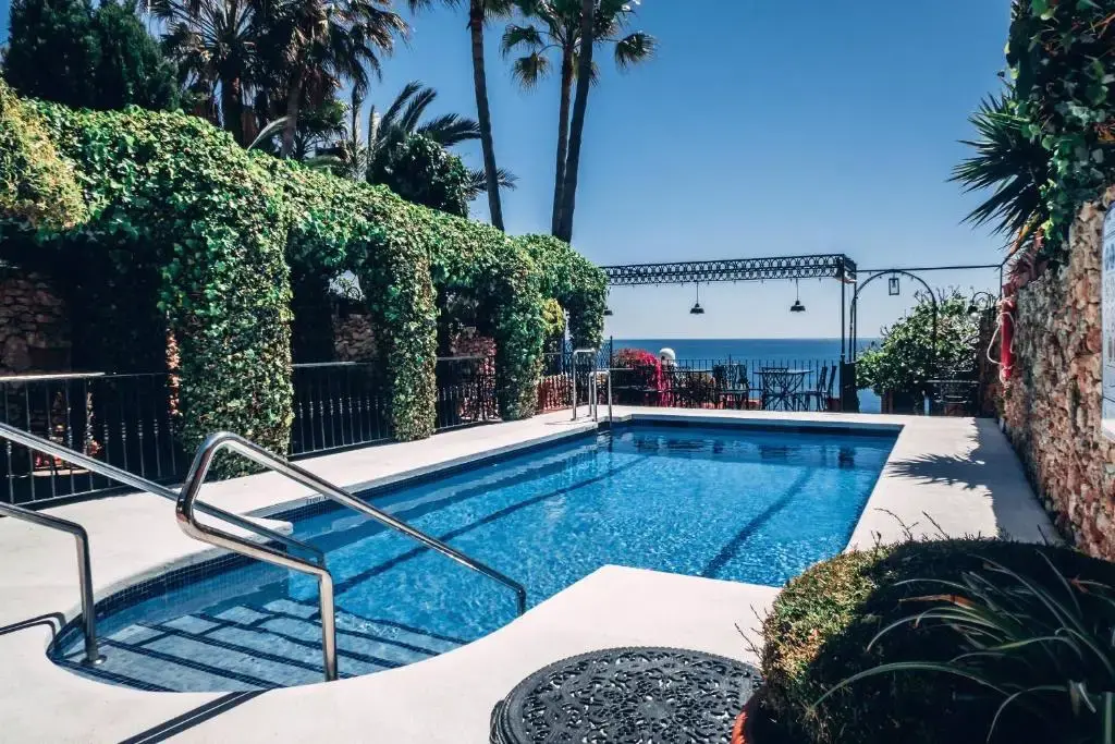 Piscina rodeada de pérgolas de hiedra y palmeras, con el mar al fondo