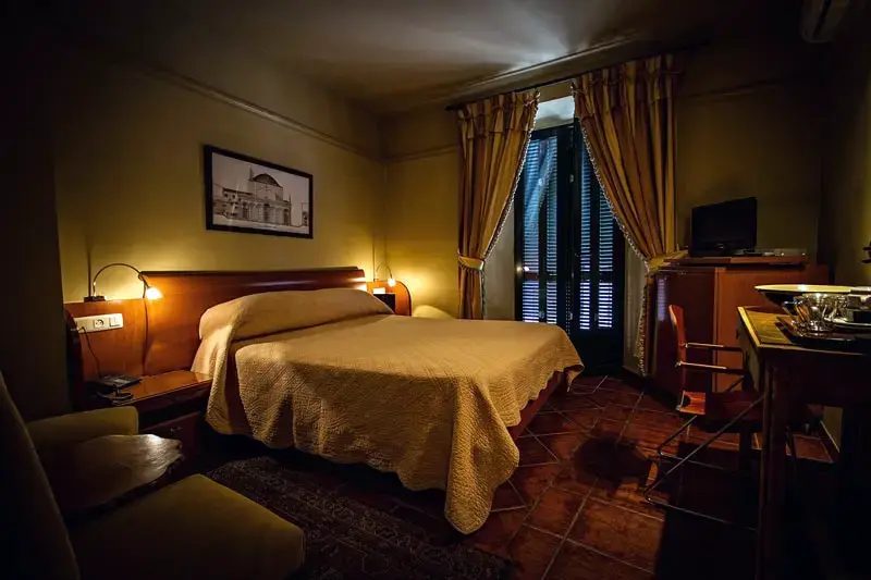 Habitación en penumbra con cama de colcha dorada, lámparas encendidas y suelo de barro