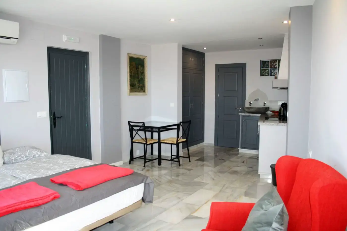 Estudio con cama, mesa con dos sillas, sillón rojo y cocina al fondo