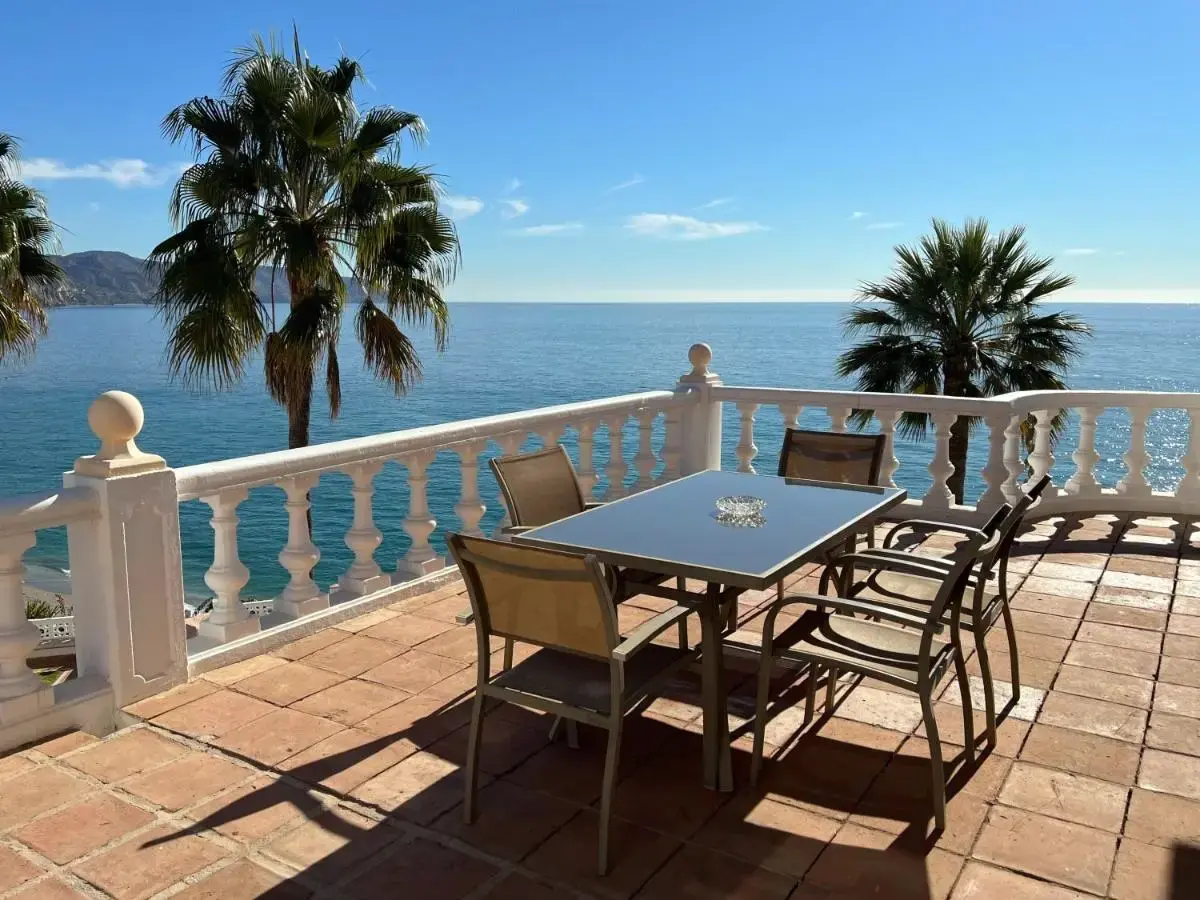 Terraza con mesa y sillas, balaustrada blanca y palmeras frente al mar en un día despejado