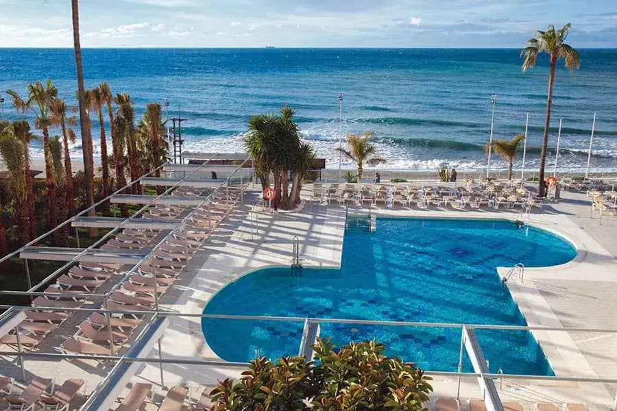 Piscina con tumbonas y palmeras junto a la playa, vista desde lo alto del hotel