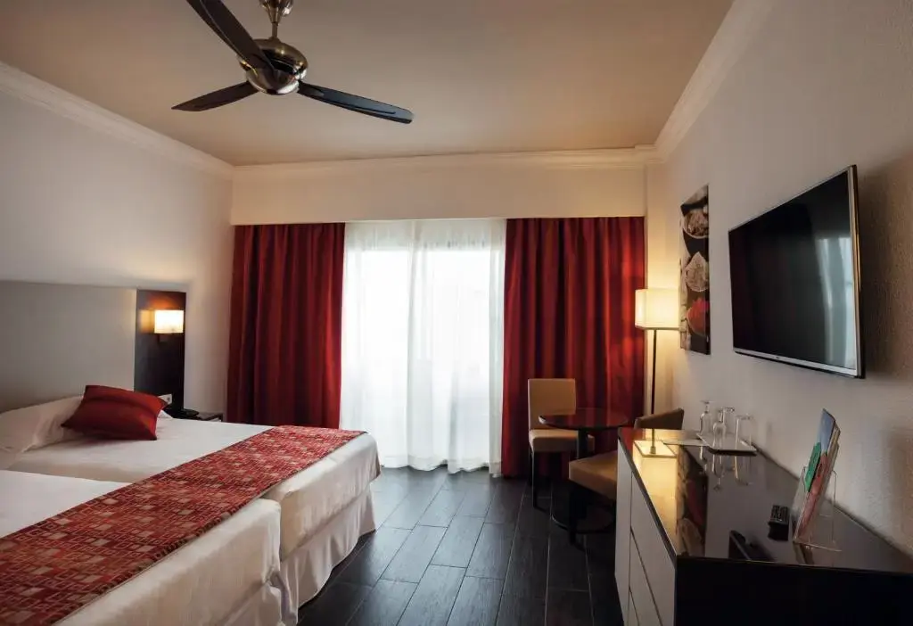Habitación con dos camas, cortinas rojas, ventilador de techo y televisor de pared