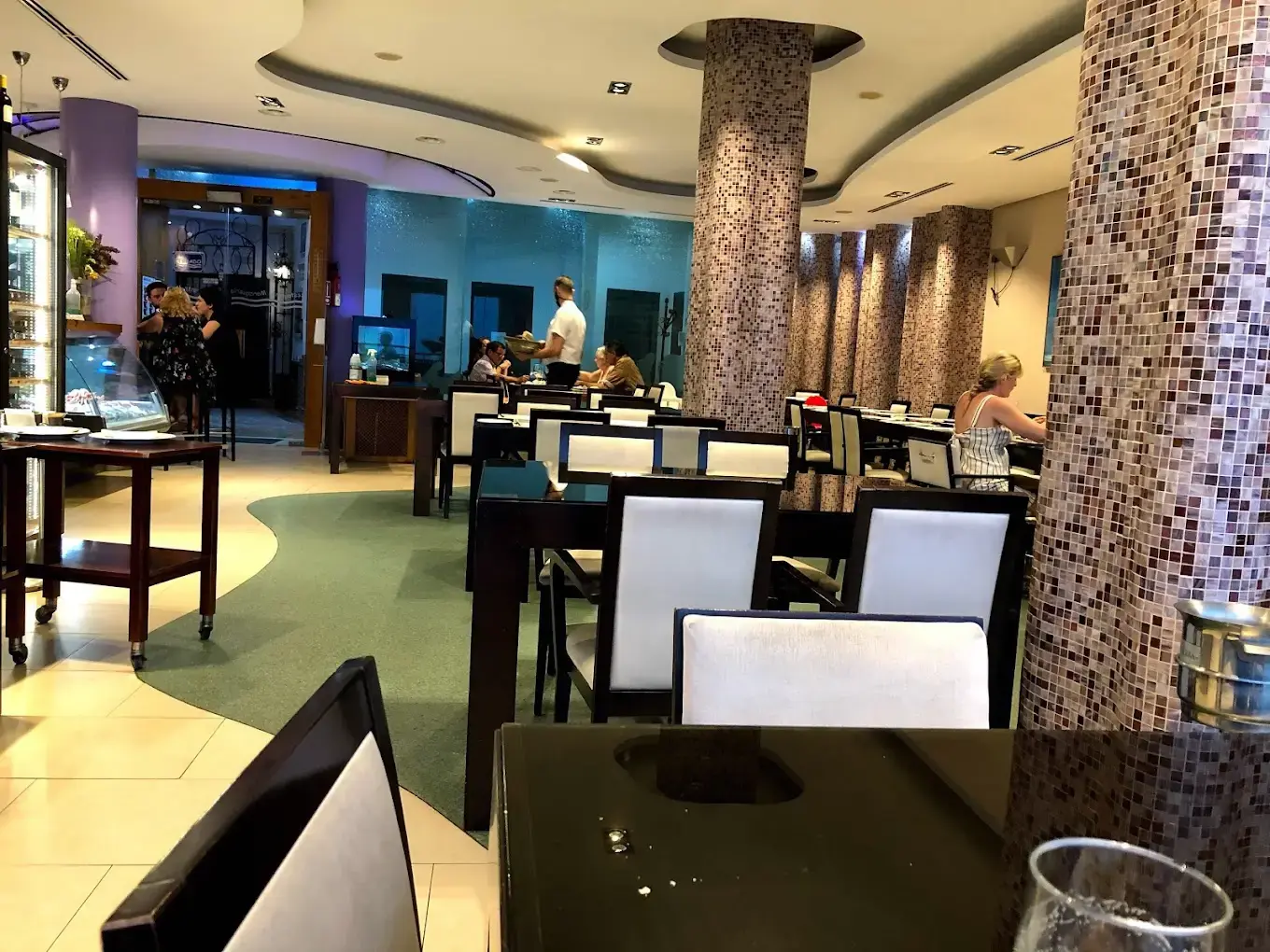 Comedor interior con columnas de mosaico, sillas blancas y vitrina junto a la entrada
