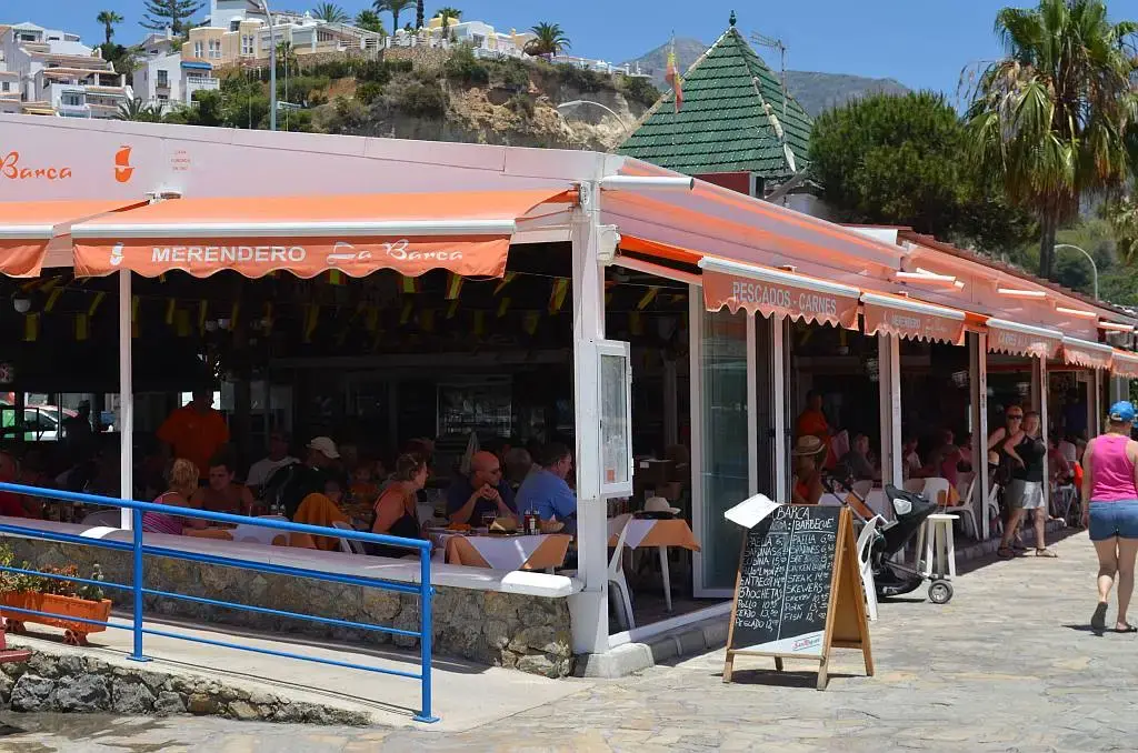 Comensales en la terraza del Merendero La Barca, con pizarra y casas en la colina