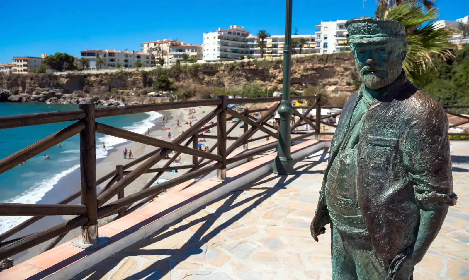 Chanquete en bronce junto a la barandilla de madera, con la playa y los edificios de Nerja