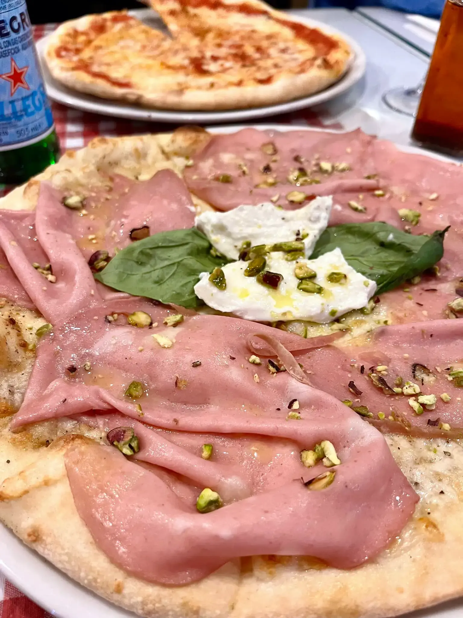Pizza de mortadela con queso fresco, pistachos y albahaca, y una margarita detrás