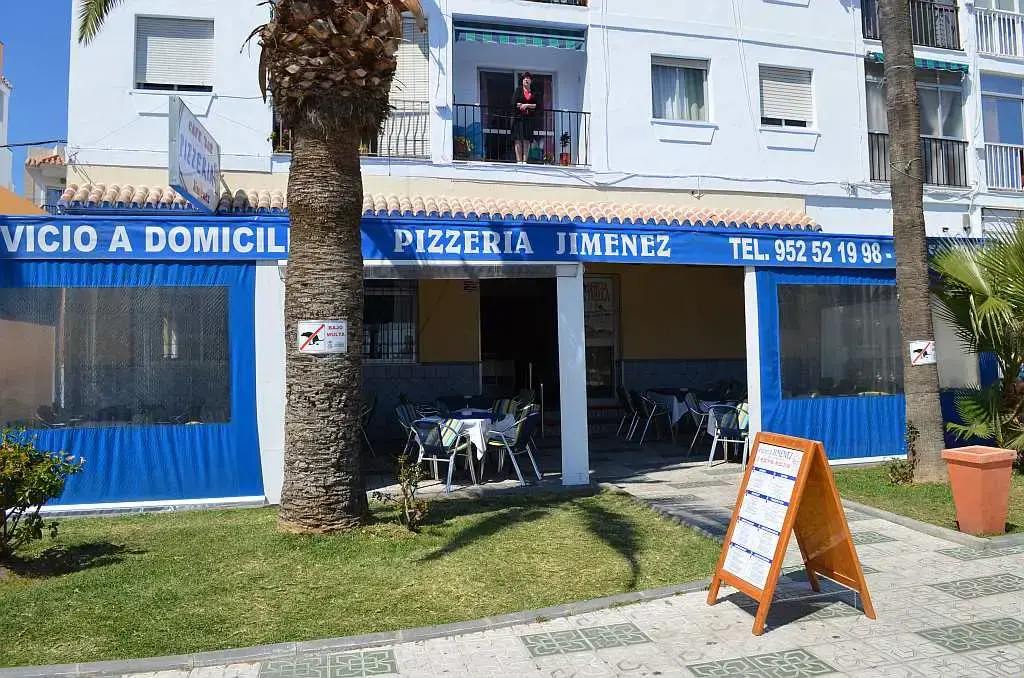 Fachada de la Pizzería Jiménez con toldo azul, servicio a domicilio y jardín delante