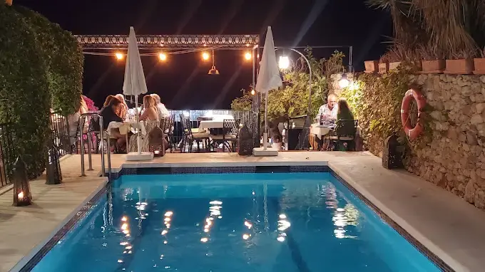 Comensales cenando junto a la piscina iluminada de noche, con guirnaldas de luces