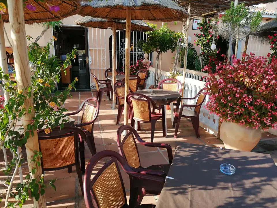Terraza soleada con sillas de rejilla, macetas de geranios y la entrada del local