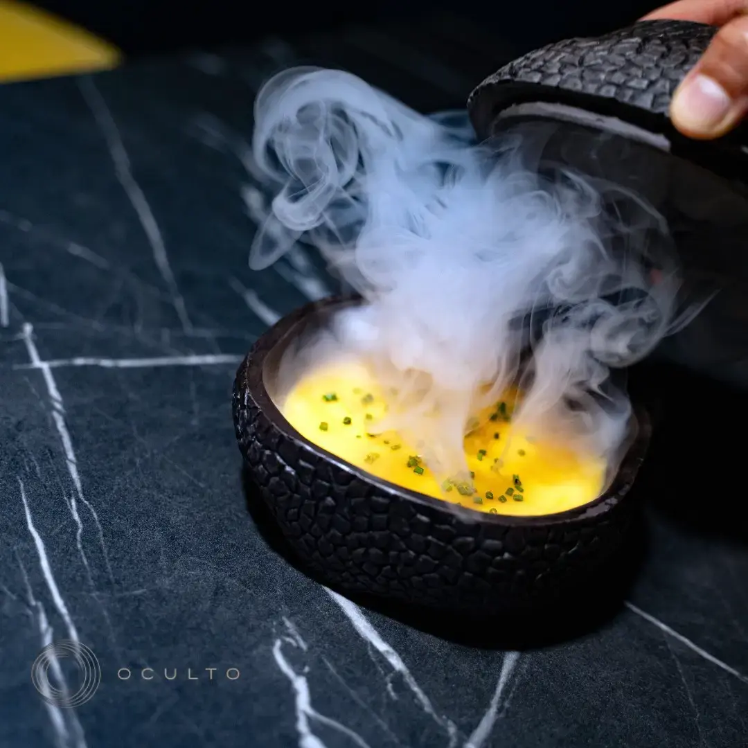 Plato servido con humo al levantar la tapa de un cuenco negro texturizado