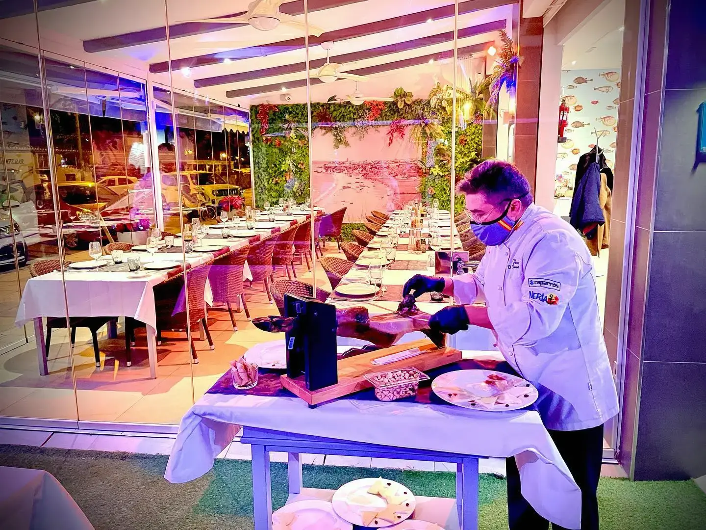 Cocinero cortando jamón a cuchillo en la terraza, con el comedor detrás
