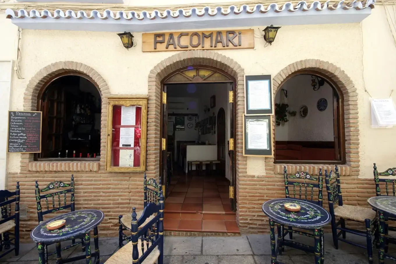 Entrada de Pacomari con arcos de ladrillo, pizarra y mesas pintadas en la acera