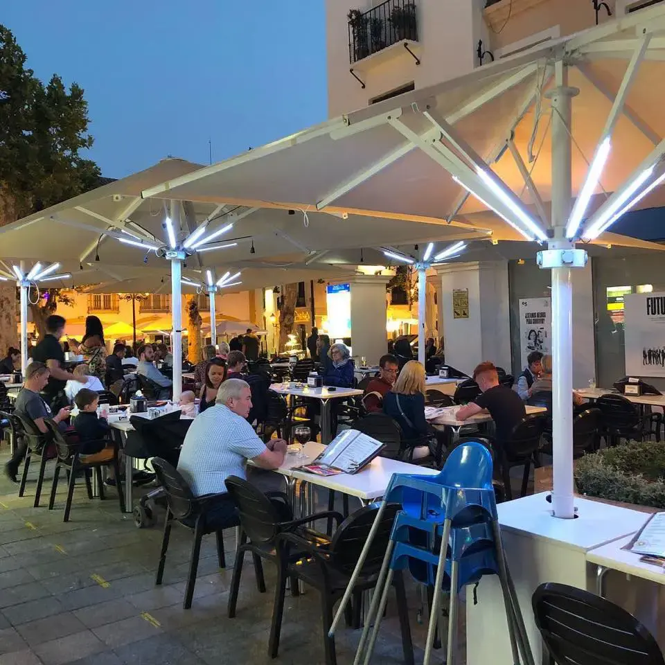Terraza llena de clientes al anochecer bajo grandes sombrillas blancas iluminadas