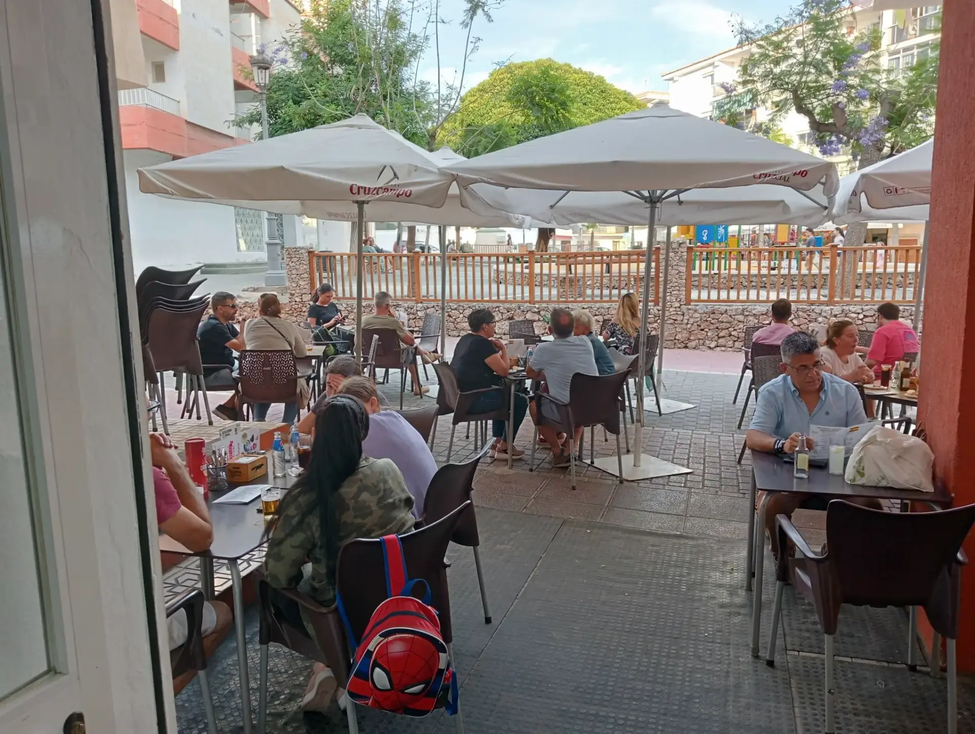 Terraza bajo grandes sombrillas abiertas, con mesas ocupadas junto a la plaza arbolada