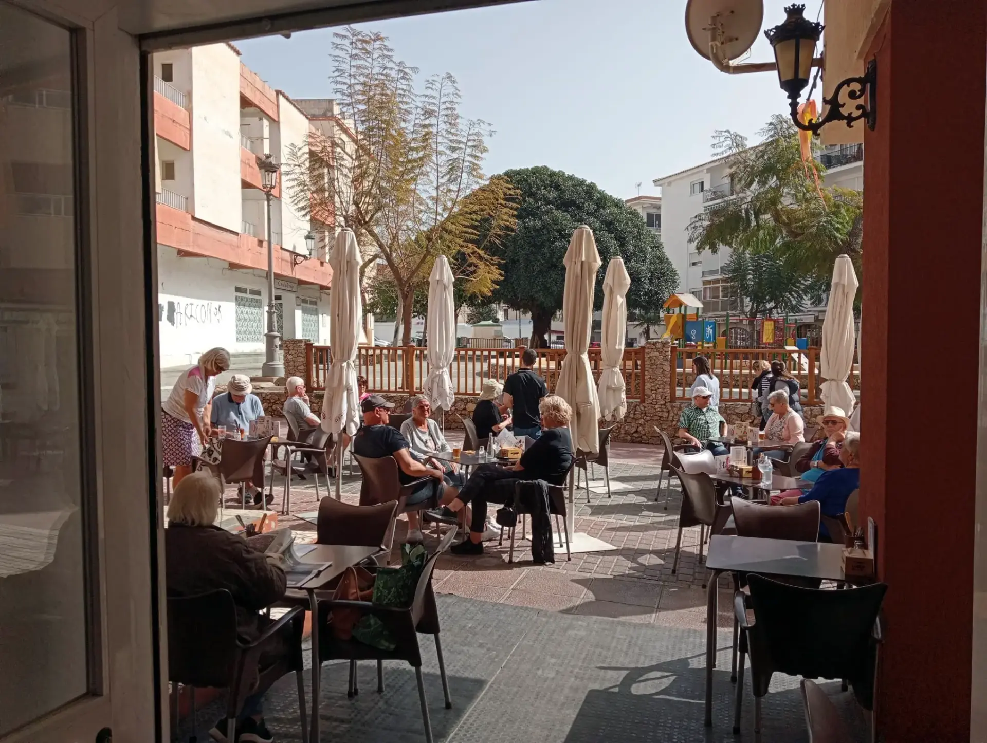 Vista de la terraza desde la puerta del bar, con árboles y un parque infantil al fondo