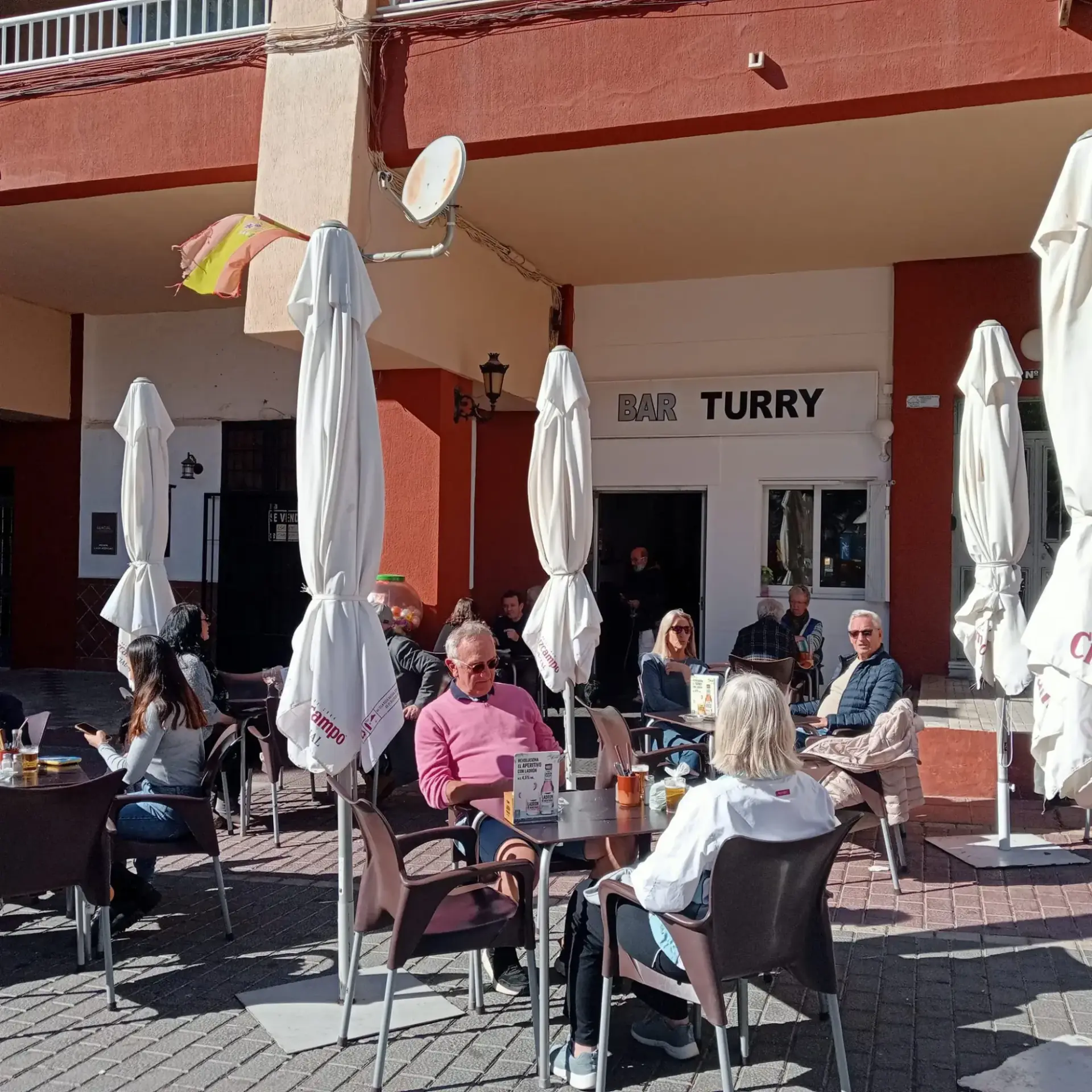 Clientes tomando algo en la terraza soleada, frente al rótulo del bar