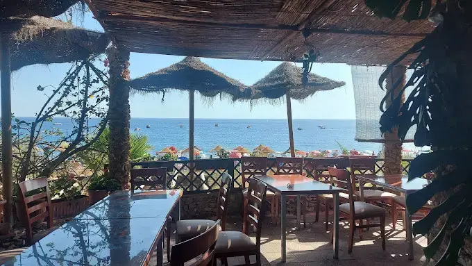 Mesas de madera bajo sombrillas de paja con vistas al mar y a la playa llena de bañistas