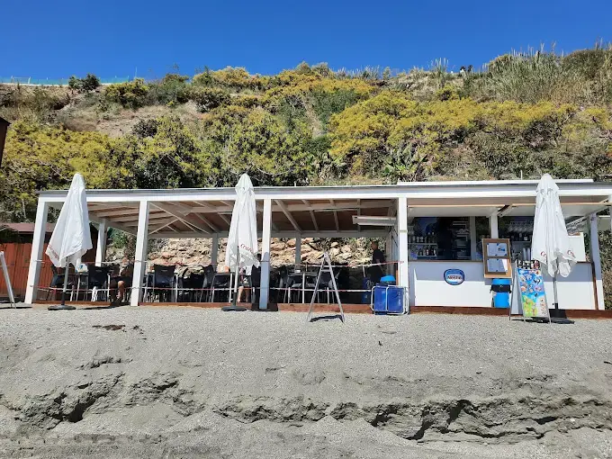 Chiringuito de madera blanca a pie de playa en Maro, bajo la ladera con vegetación