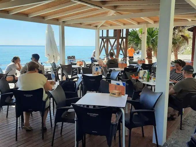 Clientes en la terraza cubierta con sillas negras y el mar azul de fondo
